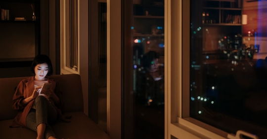 woman sitting by window at night looking at phone.