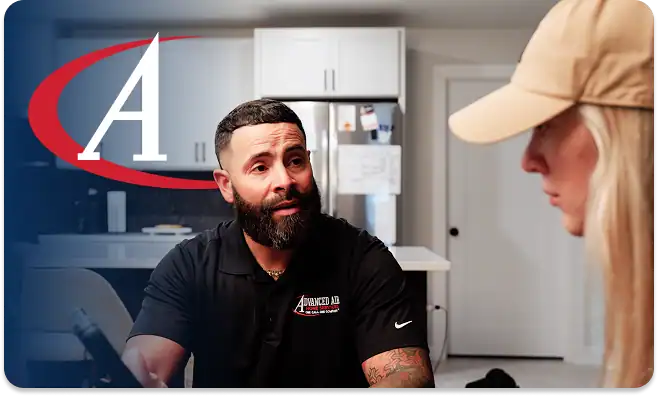A bearded man in a black polo shirt talks to a woman in a tan cap at a table in a modern kitchen, with a large “A” logo on the left side.