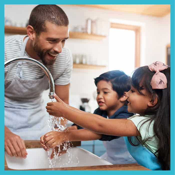 An adult helps two young children wash their hands under running water at a kitchen sink.